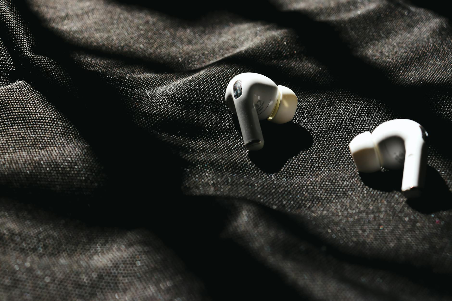 Close-up of white wireless earbuds on black fabric with dramatic sunlight and shadows.