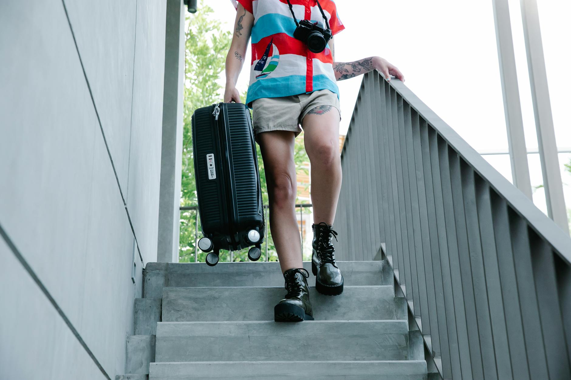 A person in casual attire holding a suitcase and camera walks downstairs.